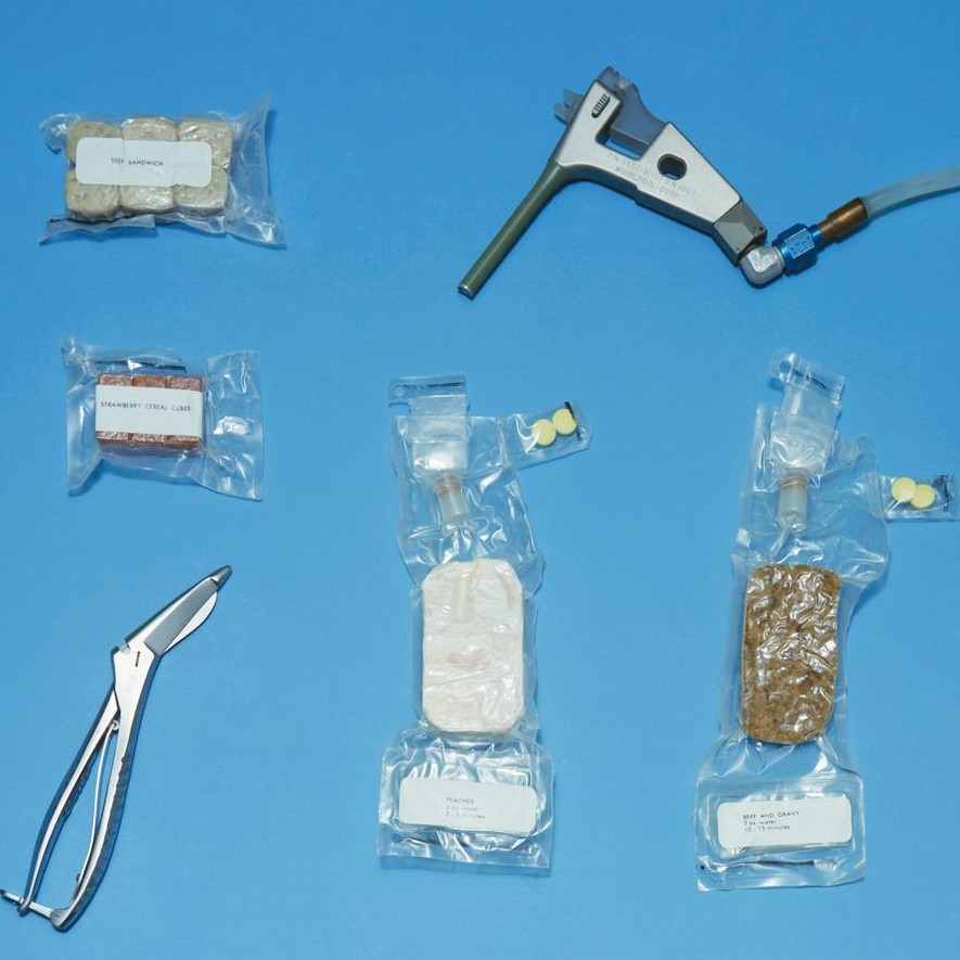 Food packages for use on the Gemini-Titan 4 (GT-4) flight. Packages include beef and gravy, peaches, strawberry cereal cubes and beef sandwiches. A water gun is used to reconstitute the dehydrated food.