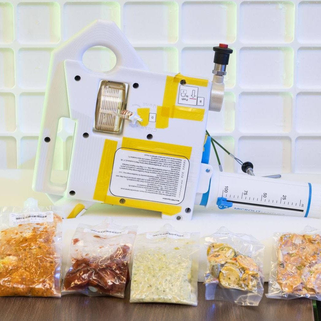 A prototype of the Mini Potable Water Dispenser, currently in development at NASA’s Marshall Space Flight Center, is displayed alongside various food pouches during a demonstration at NASA’s Johnson Space Center. 