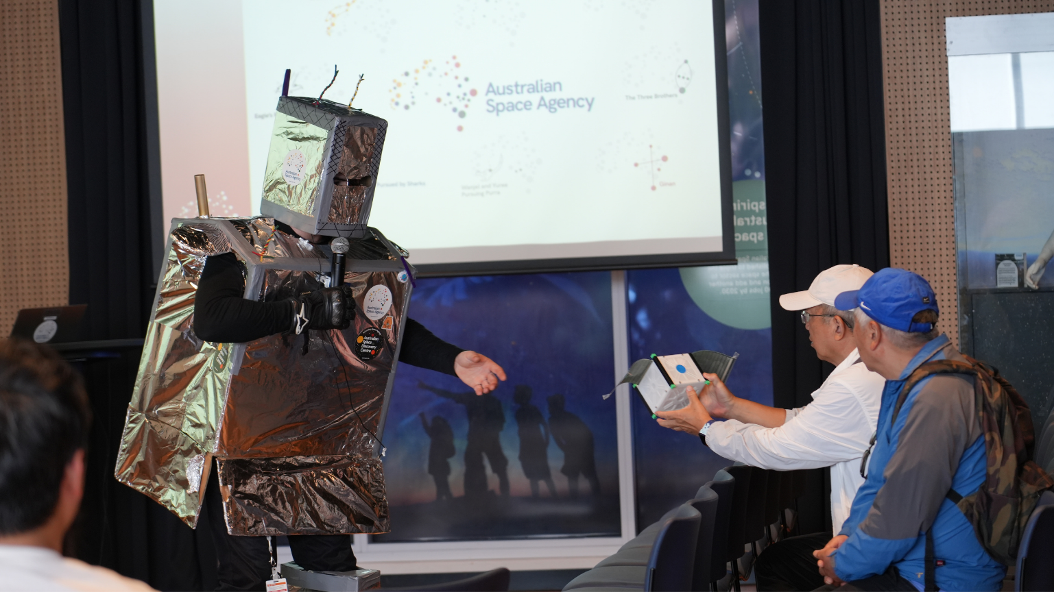 Senor space enthusiasts with a space project being presented by a person in a robot costume.