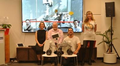 The Akin team comprising of four people who appear to be two men and two women on a couch in front of a tv screen/window with two more male appearing people and a space device behind them