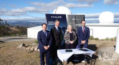 A group of three men and a woman standing behind a table with a white table cloth on it and two copies of a document in front of Thales
