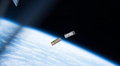 A gold and a silver CubeSat float in the dark sky of space above the Earth.