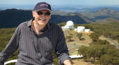Astronomer-at-large, Fred Watson, poses for a photo with the Siding Spring Observatory at a distance in the background.