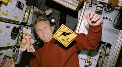 Andy Thomas floats in spaceship with stuffed kangaroo toy and kangaroos next 14km mini sign