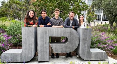 Five people in casual clothes stand behind a large 'JPL' stone monument on the ground in a garden area.