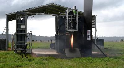 In the distance in an outdoor landscape surrounded by grass, an engine is set up on a platform and tested with a flame emerging from underneath.
