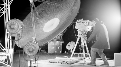 A cameraman with his equipment with satellite dish in the background.