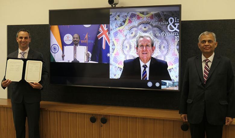 Two men standing in front of a television, with a third man on the screen. The man on the left holds the signed documents.