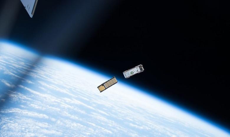 A gold and a silver CubeSat float in the dark sky of space above the Earth.