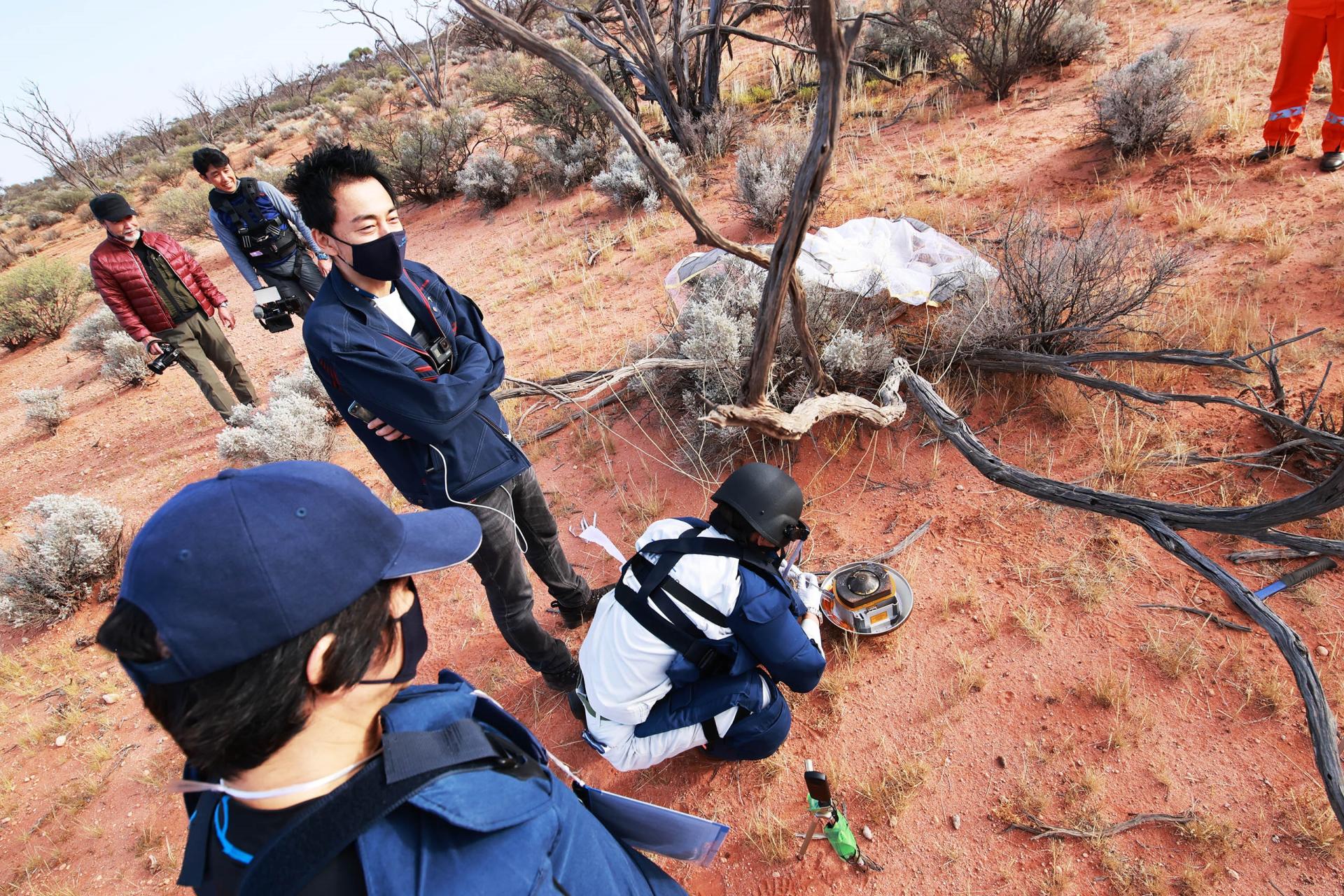 Group of people in desert. One of the people is inspecting capsule which is on the ground.